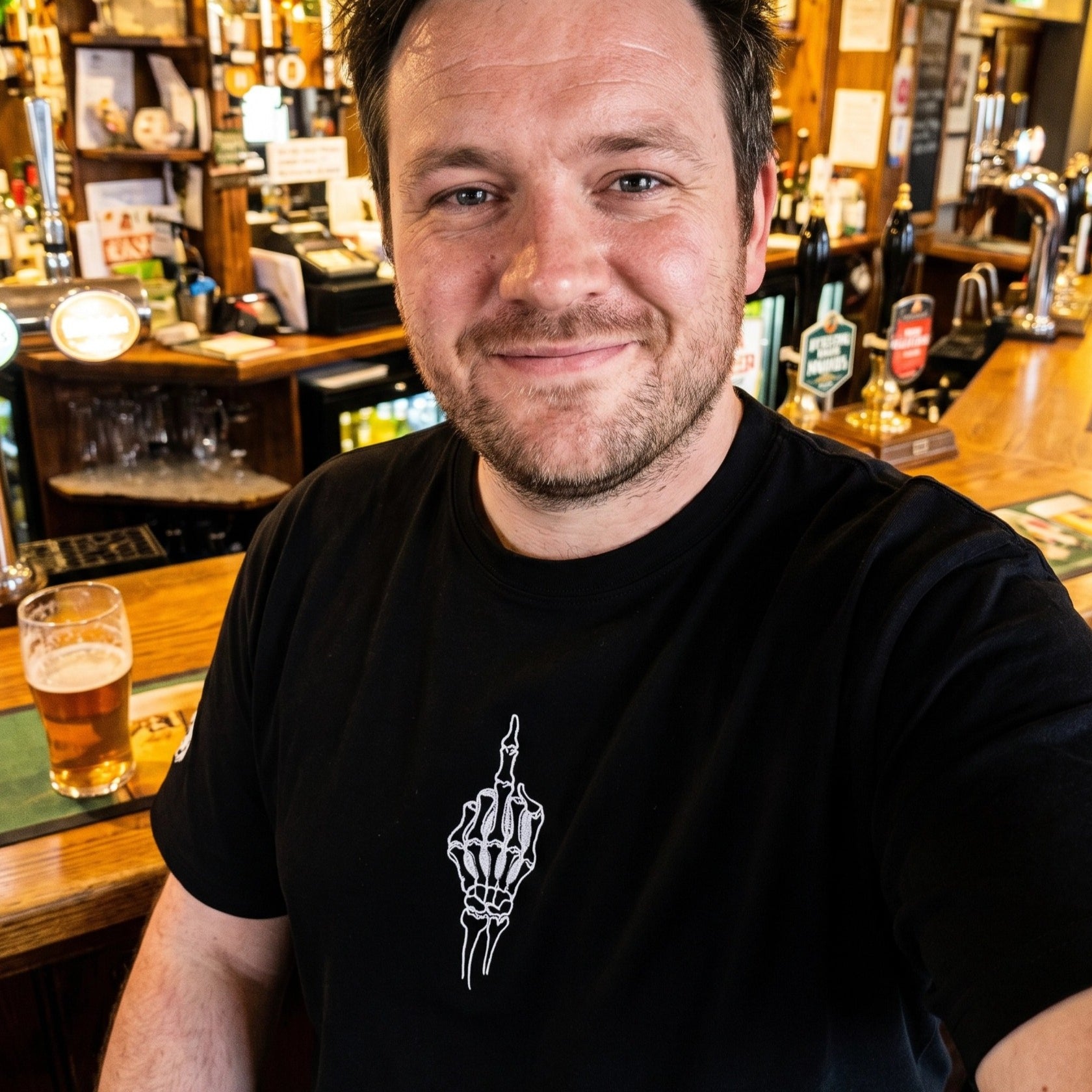 Man sitting at a bar with a beer, wearing a black t-shirt with a white graphic design.