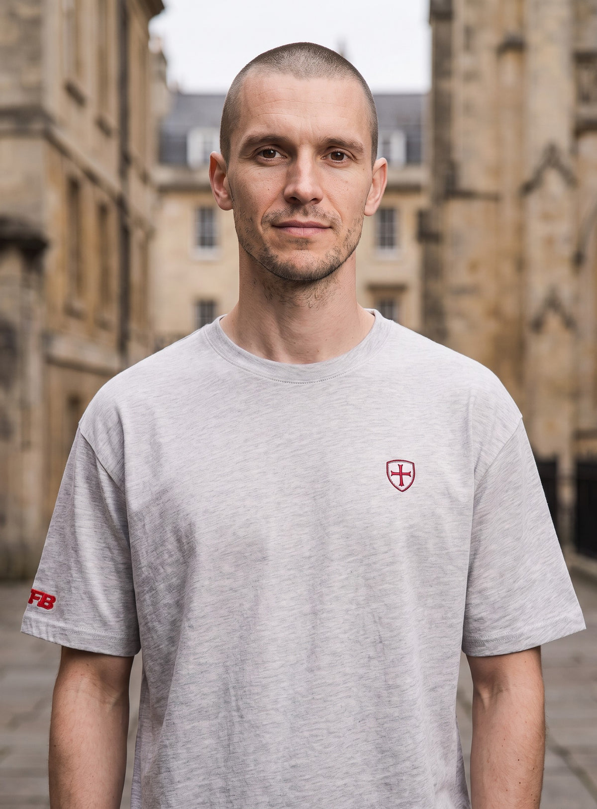 A man wearing a light gray crewneck t-shirt with a small red shield logo on the left chest, standing outdoors in front of a blurred stone building.