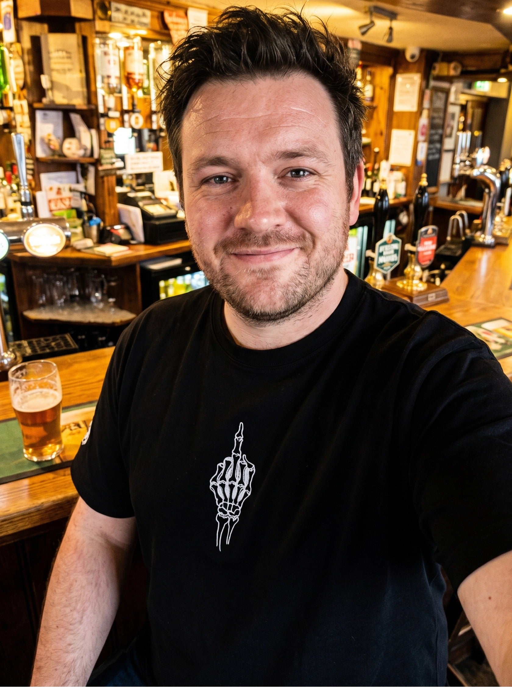 Man sitting at a bar with a beer, wearing a black t-shirt with a white graphic design.