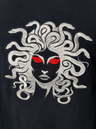 Close-up of a black garment featuring a large embroidered Medusa head graphic with white snakes for hair and bright red eyes.