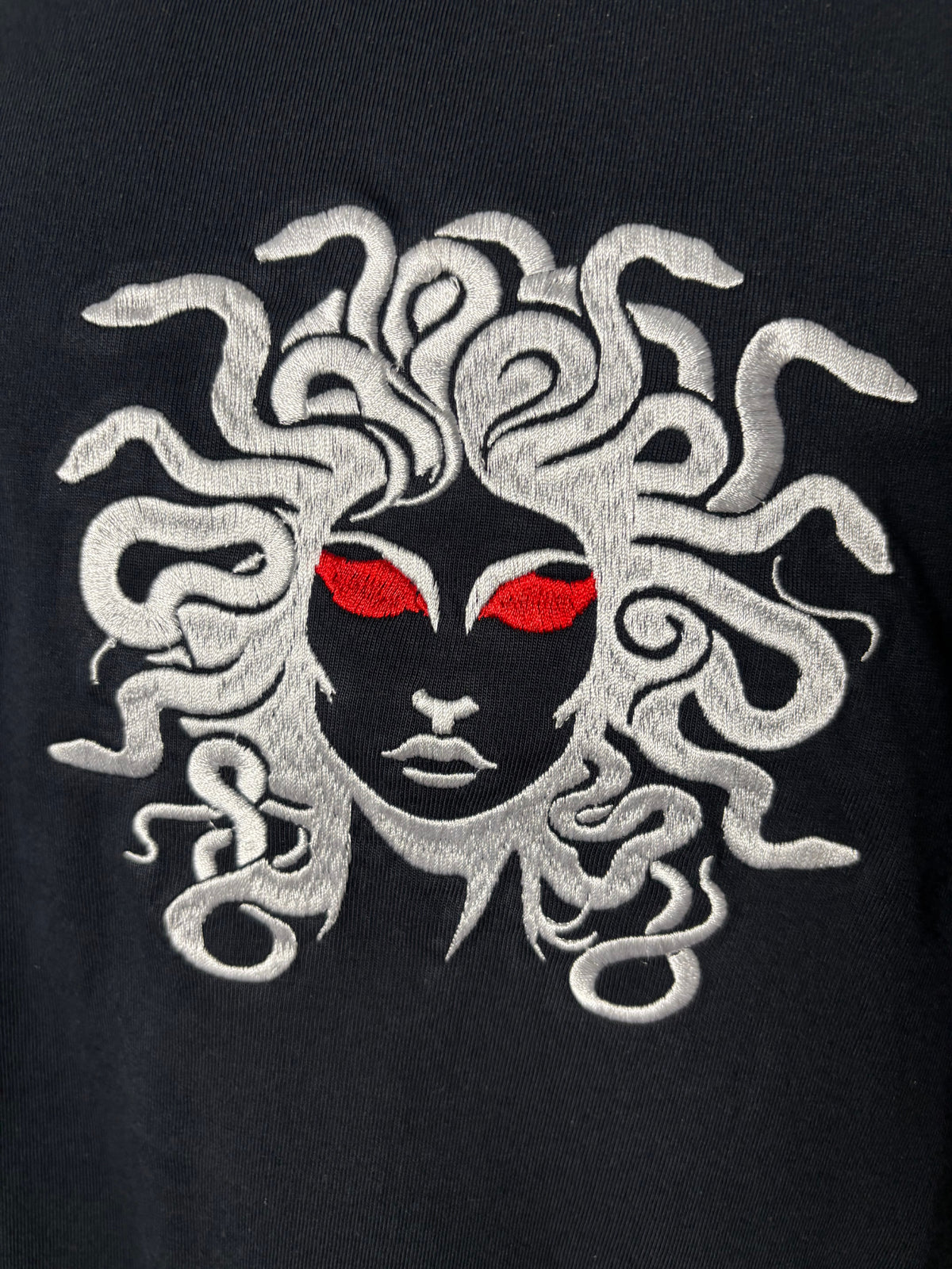 Close-up of a black garment featuring a large embroidered Medusa head graphic with white snakes for hair and bright red eyes.
